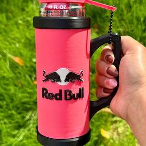 May include: A bright pink cylindrical can holder with a black handle and base, featuring the Red Bull logo and text in black. The holder contains a 355 ml energy drink can with a matching pink lid attached by a small black beaded chain.