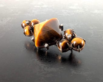 May include: A decorative brooch featuring a large, square-cut tiger's eye stone, flanked by smaller, round tiger's eye stones. The stones are set in a silver-toned metal setting, creating a striking contrast against the dark background.