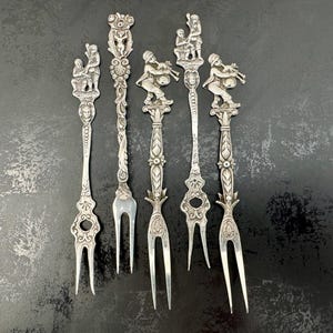 May include: A collection of antique silver serving forks. Each fork features ornate, detailed designs, including figures of musicians and other decorative elements. The forks are arranged on a dark surface, showcasing their intricate craftsmanship.