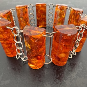 Op de afbeelding: Een vintage armband met cilindrische, amberkleurige harskralen met organische insluitsels. De kralen zijn verbonden door een zilverkleurige metalen ketting. De armband wordt gepresenteerd tegen een donkere achtergrond.