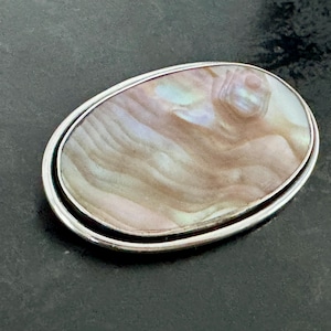 Può includere: Spilla ovale in argento con un centro perlescente e iridescente. La spilla presenta una doppia cornice argentata e un design a spirale e strutturato all'interno, che mostra sfumature di beige, rosa e blu.