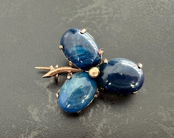 May include: A gold-toned brooch featuring three oval, deep blue gemstones arranged in a cloverleaf pattern. The brooch has a delicate, branch-like design with a central gold bead.