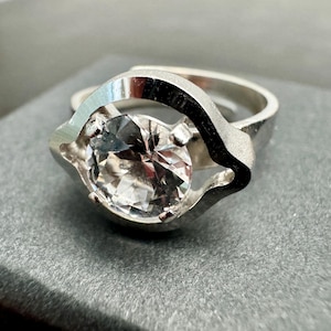 May include: A silver-toned ring featuring a large, round, clear faceted stone set within a smooth, asymmetrical metal frame. The band is open at the back, and the metal surface displays a polished finish with reflective highlights throughout the design.