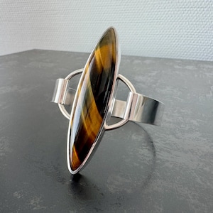 May include: A silver ring featuring a large, oval tiger's eye stone. The stone is a mix of brown, gold, and black. The ring has a modern design with a wide band and a circular accent that holds the stone.