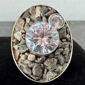Può includere: Anello d'argento ovale con una grande gemma rotonda e trasparente e una più piccola. L'incastonatura è riempita di piccole pietre grigie e beige. L'anello è presentato su uno sfondo nero.