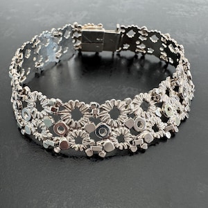 May include: A silver-toned metal bracelet featuring an intricate, openwork design composed of repeating circular, flower-like motifs and small geometric shapes. The bracelet includes a rectangular box clasp closure at the top, creating a continuous band.