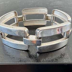 May include: A silver-coloured bracelet with a linked design. The bracelet features rectangular links that are connected to each other. The metal has a brushed finish, giving it a matte appearance. The bracelet is displayed on a dark surface.