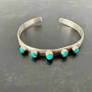 May include: Silver cuff bracelet with five round turquoise stones. The bracelet has a textured design along the band and is open at the ends. The turquoise stones are set in silver bezels.