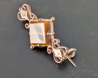 May include: A vintage gold-toned brooch with a rectangular amber-colored stone at its center. The brooch features ornate scrollwork and decorative elements, with a pin closure on the back. The design is symmetrical and elegant.