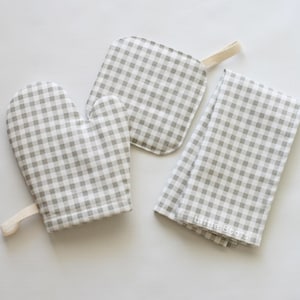 May include: A kitchen set featuring a gray and white gingham pattern. The set includes an oven mitt, a pot holder, and a folded kitchen towel. The towel has the text "Little Bears" printed on it. The items are made of fabric.