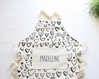 May include: A white apron with black hand-drawn hearts and a large pocket with the name "Madeleine" printed on it.