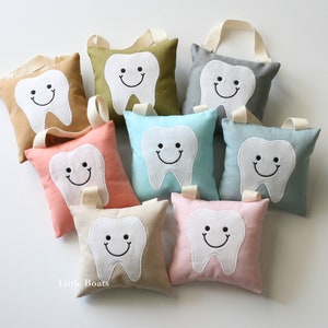 May include: Eight tooth-shaped pillows with smiling faces, each in a different color: tan, green, gray, light blue, pink, beige, and light pink. The pillows have a loop for hanging. The text "Little Boats" is visible in the bottom left corner.