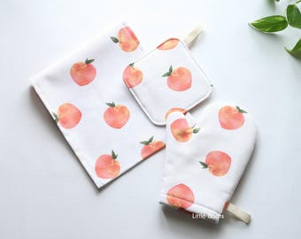 May include: A white kitchen set featuring a peach pattern. The set includes a rectangular towel, a square pot holder, and an oven mitt. Each item is decorated with watercolor-style peaches and green leaves.