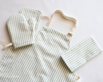 May include: A child's apron, oven mitt, and pot holder set. The set features a white background with vertical green stripes. The apron has a beige neck strap and waist ties. The oven mitt and pot holder are also striped.