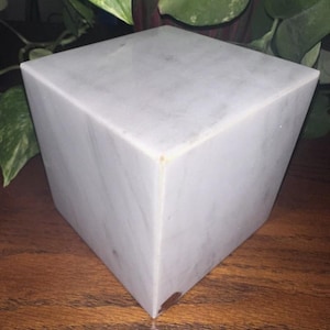 May include: A white marble cube with a smooth, polished surface. The cube is sitting on a wooden surface.