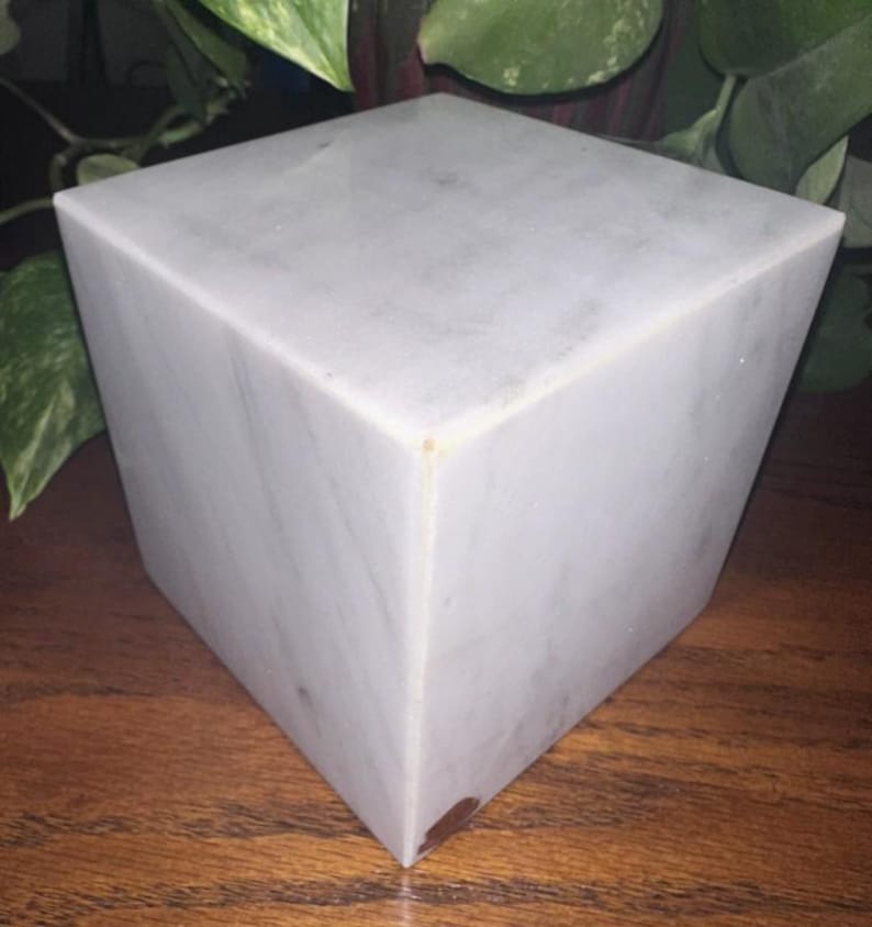 May include: A white marble cube with a smooth, polished surface. The cube is sitting on a wooden surface.
