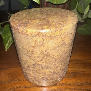 May include: A round, brown and yellow stone container with a lid. The container has a speckled pattern and a smooth, polished finish.