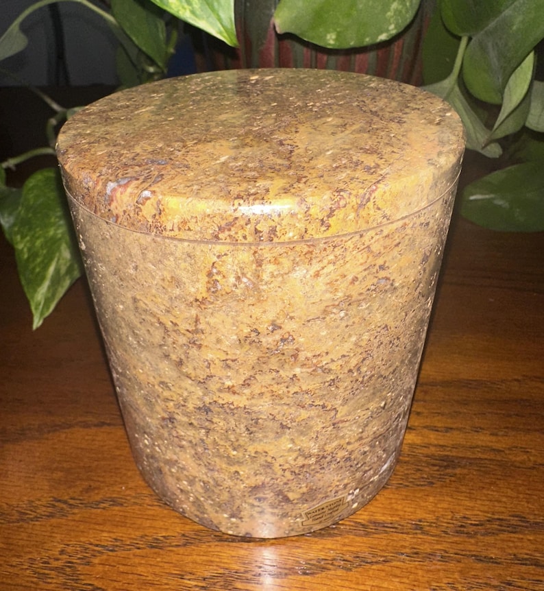 May include: A round, brown and yellow stone container with a lid. The container has a speckled pattern and a smooth, polished finish.