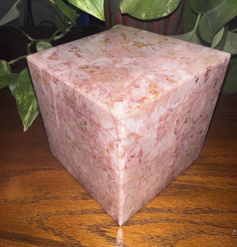 May include: A pink marble cube box with a light pink and white marbled pattern. The box has a smooth, polished surface.