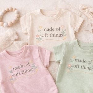 May include: Three pastel-coloured baby t-shirts, cream, pink, and mint green, each printed with the words "made of soft things" in a floral design. A wooden bead bracelet and other baby accessories are visible.