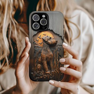May include: A phone case with a detailed relief of a dog, wheat, and a sunset. The case has a wood-grain texture and a dark border. The dog is brown, and the sunset is orange. The case is held by a person.