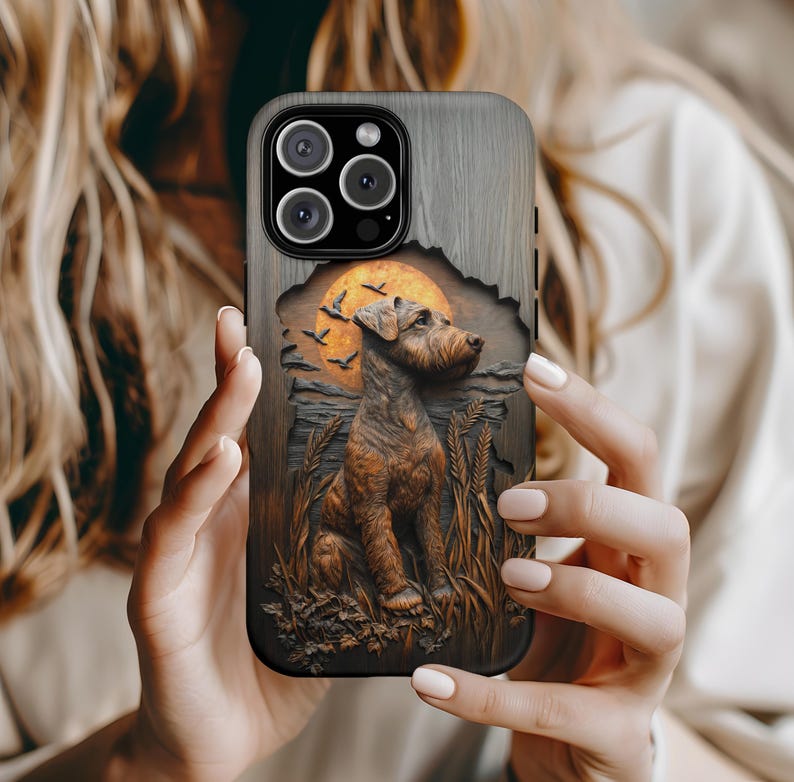 May include: A phone case with a detailed relief of a dog, wheat, and a sunset. The case has a wood-grain texture and a dark border. The dog is brown, and the sunset is orange. The case is held by a person.