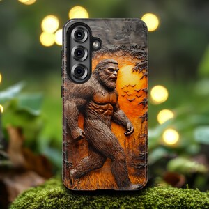May include: A phone case featuring a detailed relief of Bigfoot walking through a forest at sunset. The case is brown with a black border and has a three-lens camera cutout.