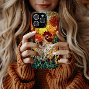 May include: A phone case featuring a colorful rooster design. The case has a vibrant stained-glass style with a rooster, trees, and a sunset. The case is held in front of a person wearing a brown sweater.