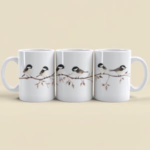 May include: Set of three white ceramic mugs with a black and white watercolor design of birds on a branch. The birds are facing left and right, and the branch has brown leaves.