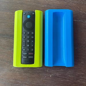 Op de afbeelding: Een limoengroene en blauwe afstandsbedieninghouder. De limoengroene houder omsluit een zwarte Fire TV-afstandsbediening, terwijl een aparte blauwe houder ernaast ligt. De afstandsbediening heeft verschillende knoppen en het Fire TV-logo.