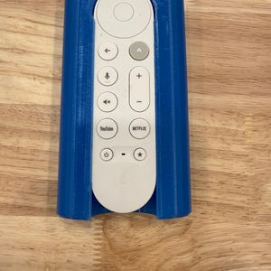 Może przedstawiać: Niebieski, drukowany w 3D uchwyt na pilota. Uchwyt otacza biały pilot z przyciskami nawigacji, głośności i usług strumieniowania, takich jak YouTube i Netflix. Uchwyt został zaprojektowany tak, aby bezpiecznie trzymać pilota.