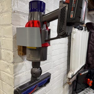 Puede incluir: Una aspiradora inalámbrica Dyson montada en una pared de ladrillos blancos. La aspiradora tiene una carcasa de motor azul y roja, un mango gris y un cabezal de limpieza negro. Una batería negra está sujeta al mango.