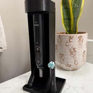 Puede incluir: Un soporte negro para cepillo de dientes con un cepillo de dientes eléctrico y un cabezal de repuesto. El soporte está sobre una encimera blanca junto a una planta en maceta con hojas verdes y amarillas. El cepillo de dientes tiene la marca "Oral-B".
