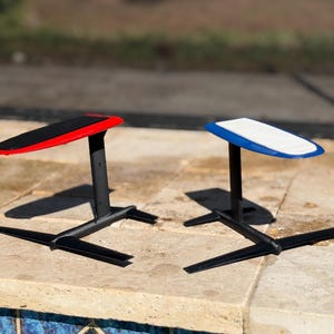 May include: Two hydrofoil boards with black supports and wings. One board has a red edge and a black top, the other has a blue edge and a white top. The boards are designed for water sports.