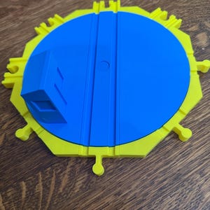 May include: A blue and yellow plastic toy train turntable. The turntable has a blue circular centre with two parallel tracks and a yellow outer ring with connector tabs. A blue rectangular block is attached to the side.
