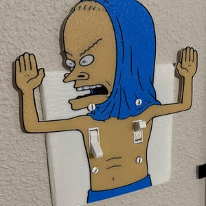 May include: A light switch cover featuring a glittery tan cartoon character with a blue hood and blue shorts, depicted with arms raised and hands open. The character has a wide-eyed expression and is integrated with two white toggle switches.