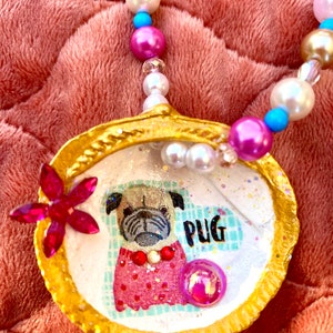 May include: A gold-colored seashell pendant necklace with a white background featuring a pug wearing a red and white polka dot dress. The word "PUG" is written in black letters on the seashell. The necklace is made with white, pink, and turquoise beads.