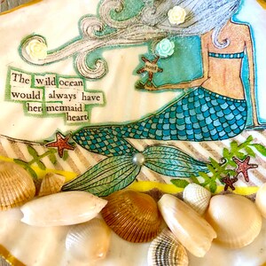 May include: A white seashell with a gold trim, decorated with a mermaid illustration, seashells, and the text "The wild ocean would always have her mermaid heart".