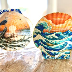 May include: Two decorative scallop shells featuring hand-painted scenes. The left shell depicts a seagull perched on driftwood at sunset. The right shell displays a stylized blue ocean wave against an orange sunset sky, both accented with small shells.