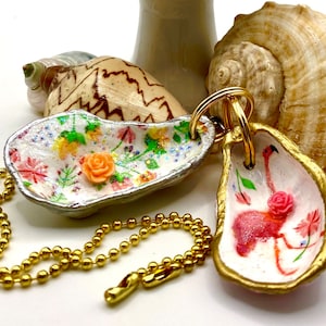 May include: Two oyster shell keychains with gold trim. One features a pink flamingo with a pink rose and the other has a floral design with a peach rose. Both are attached to a gold chain.