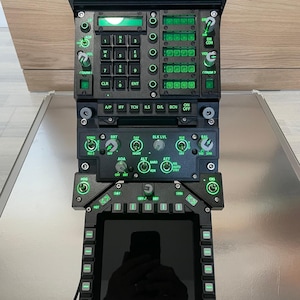 Puede incluir: Un panel de simulador de vuelo negro y verde con varios botones, perillas y una pequeña pantalla. El panel tiene etiquetas como "ADF", "VOL", "COMM", "CLR", "ENT", "A/P", "IFF", "TCN", "ILS", "D/L", "BCN", "ON", "OFF", "EM", "CON", "COMM2", "NORM", "BRT", "DAY", "BLK LVL", "BAL", "REJ2", "OFF", "NIGHT", "VID SYM", "AOA", "ALT", "ATT", "BARO", "INS", "AUTO", "ROR", "STBY", "OFF SRT", "HOG", "CRS", "DAY", "SYM", "OFF BRT", "GAIN", "CONT".