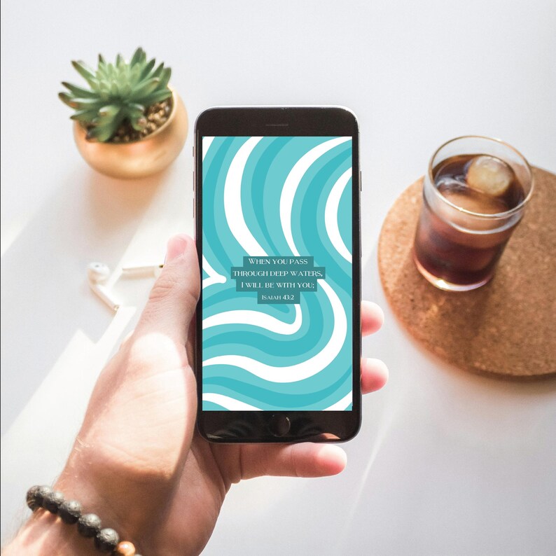 Puede incluir: Un tel&eacute;fono inteligente muestra un patr&oacute;n ondulado turquesa y blanco con el texto "WHEN YOU PASS THROUGH DEEP WATERS, I WILL BE WITH YOU; ISAIAH 43:2." Tambi&eacute;n se ven una peque&ntilde;a suculenta en una maceta dorada, un vaso de bebida helada y auriculares.