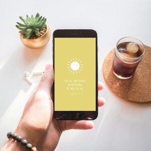 Puede incluir: Un smartphone negro muestra una pantalla amarilla con un sol blanco y el texto: "Echando toda vuestra ansiedad sobre &eacute;l, porque &eacute;l se preocupa por vosotros." Tambi&eacute;n se ve una peque&ntilde;a suculenta en una maceta dorada y un vaso de bebida helada.