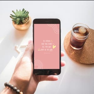 Puede incluir: Un tel&eacute;fono inteligente muestra una pantalla rosa con el texto "BE STRONG AND TAKE HEART ALL YOU WHO HOPE IN THE LORD." El tel&eacute;fono se sostiene en una mano, con una planta suculenta y un vaso de bebida helada en el fondo.