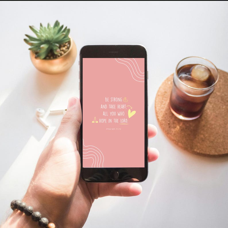 Puede incluir: Un tel&eacute;fono inteligente muestra una pantalla rosa con el texto "BE STRONG AND TAKE HEART ALL YOU WHO HOPE IN THE LORD." El tel&eacute;fono se sostiene en una mano, con una planta suculenta y un vaso de bebida helada en el fondo.