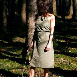 Könnte beinhalten: Eine Frau in einem hellgrünen, ärmellosen Kleid steht in einem Wald. Das Kleid hat ein schlichtes Design und ist aus einem weichen, fließenden Stoff gefertigt.