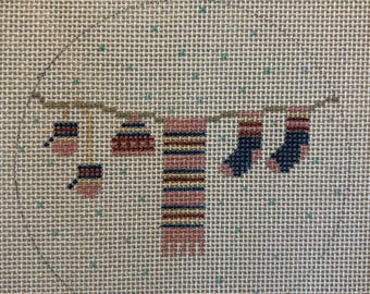 May include: Needlepoint design of a clothesline with hanging items. The items include a pink and blue striped scarf, two pairs of socks, and two small items. The background is a beige canvas with small green and blue dots.