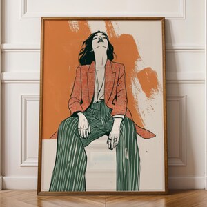 May include: A framed art print featuring a stylized illustration of a person in a red blazer and green striped pants. The artwork has an orange and cream background and is displayed against a white wall and wooden floor.