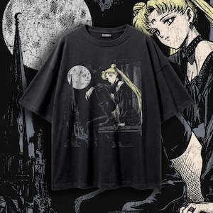 Puede incluir: Una camiseta negra de manga corta con un estampado gráfico de un personaje rubio de pelo largo sentado en una repisa frente a una luna llena y un fondo arquitectónico. El diseño tiene un aspecto desgastado y texturizado.