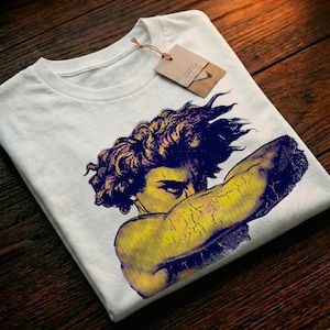 Puede incluir: Camiseta blanca con un gráfico estilizado de una persona con piel amarilla y cabello y brazo azul. La persona se cubre la cara con el brazo. Una etiqueta marrón está adherida a la camiseta.